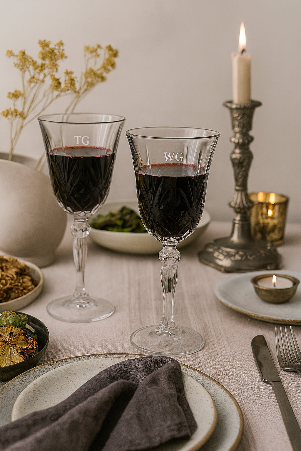 Two engraved William wine glasses with initials on a table setting with candles and plates.