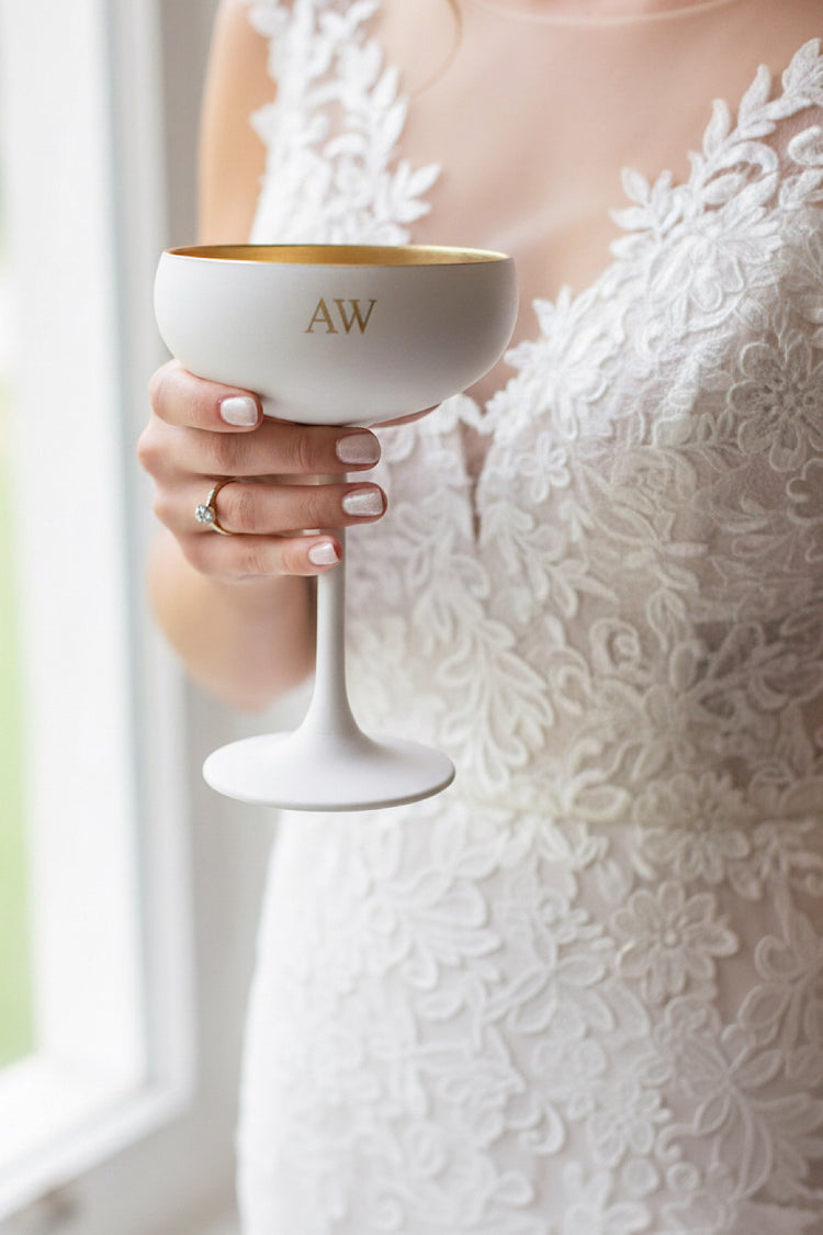 Bride holding a white champagne saucer glass with gold rim and initials 'AW' in a blurred background.